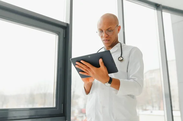 Mature african doctor using digital tablet in corridor . Portrait of confident male doctor using tablet computer in clinic with copy space. Successful smiling doctor in labcoat wearing stethoscope.