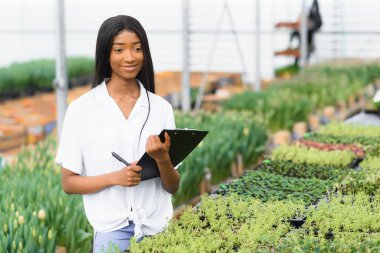 Tarım yönetimi. Gülümseyen Afrikalı Amerikalı kız, seradaki, yan manzaradaki, serbest alandaki çiçeklerin fotoğrafını çekiyor.