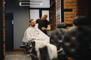 Genç yakışıklı berber berber dükkanında sakallı çekici bir adamın saçını kesiyor.