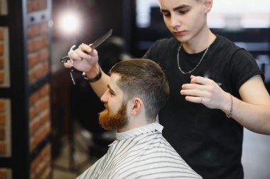 Saç kesimini mükemmel gösteriyor. Genç sakallı adam berber dükkanında sandalyede otururken kuaför tarafından tıraş oluyor.