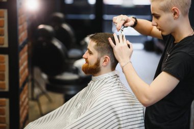 Saç kesimini mükemmel gösteriyor. Genç sakallı adam berber dükkanında sandalyede otururken kuaför tarafından tıraş oluyor.