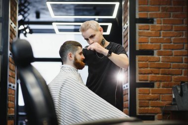 Saç kesimini mükemmel gösteriyor. Genç sakallı adam berber dükkanında sandalyede otururken kuaför tarafından tıraş oluyor.