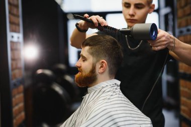 Genç yakışıklı berber berber dükkanında sakallı çekici bir adamın saçını kesiyor.