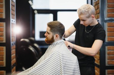 Saç kesimini mükemmel gösteriyor. Genç sakallı adam berber dükkanında sandalyede otururken kuaför tarafından tıraş oluyor.