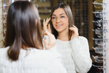Kadın dükkanda bardak seçiyor. Beyaz kazaklı esmer gözlük alır. Farklı gözlük modelleri olan arka plandaki kız..