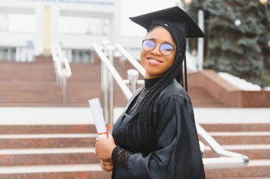Mezuniyet şapkası ve cübbesi içinde okul binasının önünde güzel bir Afrikalı üniversite öğrencisi.