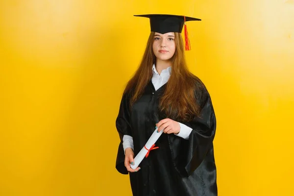 Happy attractive graduate in a master's dress, with a diploma on a yellow background. Concept of the graduation ceremony