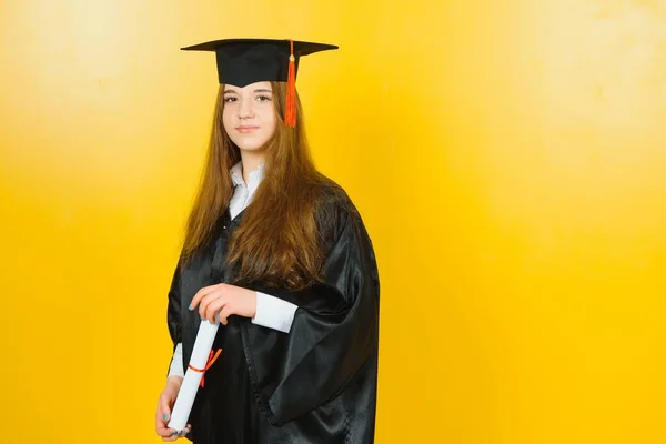Happy attractive graduate in a master\'s dress, with a diploma on a yellow background. Concept of the graduation ceremony