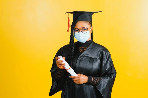 Portrait of a young african student in a graduate cap protective mask, on a yellow background, Graduation 2021