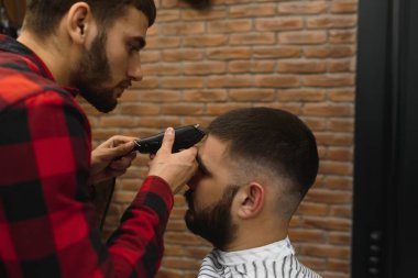 Saç kesimini mükemmel gösteriyor. Genç sakallı adam berber dükkanında sandalyede otururken kuaför tarafından tıraş oluyor.