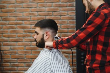 Saç kesimini mükemmel gösteriyor. Genç sakallı adam berber dükkanında sandalyede otururken kuaför tarafından tıraş oluyor.