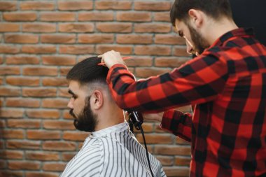 Saç kesimini mükemmel gösteriyor. Genç sakallı adam berber dükkanında sandalyede otururken kuaför tarafından tıraş oluyor.