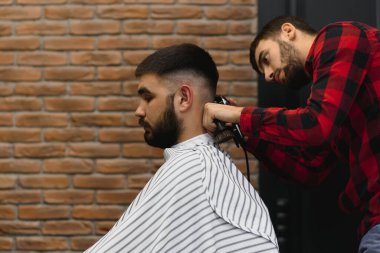 Yakışıklı sakallı adam kuaförde saçını kestirirken gülümsüyor.