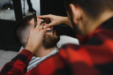 Berber dükkanında berber koltuğunda otururken tıraş makinesiyle sakal tıraşı olan ciddi sakallı adam. Berber Dükkanı Teması