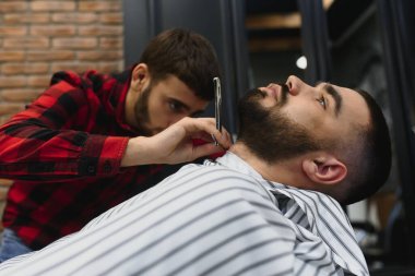 Berber dükkanında berber koltuğunda otururken tıraş makinesiyle sakal tıraşı olan ciddi sakallı adam. Berber Dükkanı Teması