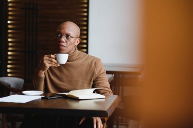 Bilgi, öğrenme, eğitim ve teknoloji. Gayri resmi giyinmiş, Afro saç stili çözen, matematiksel problemler çözen, kafeterya masasında ders kitapları ve elektronik cihazlarla oturan bir öğrenci.