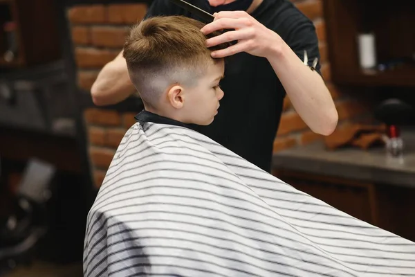 Children hairdresser with scissors is cutting little boy against a dark background. Contented cute preschooler boy getting the haircut.