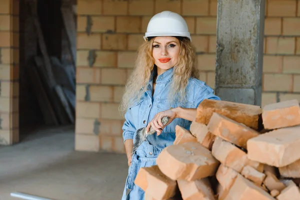 Attractive female construction worker in hardhat. Confident young specialist in checkered blue shirt in jeans standing in empty room. Interior design and renovation service.