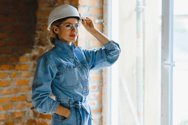 Attractive female construction worker in hardhat. Confident young specialist in checkered blue shirt in jeans standing in empty room. Interior design and renovation service.