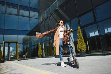Ceketli genç ve güzel bir kadın ofis binalarında çalışırken gülümsüyor ve elektrikli scooter kullanıyor.