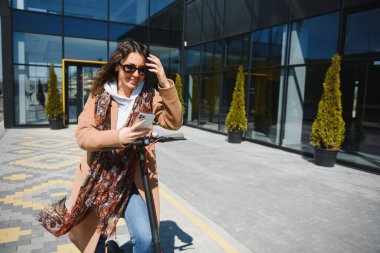Genç ve güzel bir kız elektrikli scooterının yanında duruyor. Gülümsüyor, iş eğitimli, arka planda modern mimari var..
