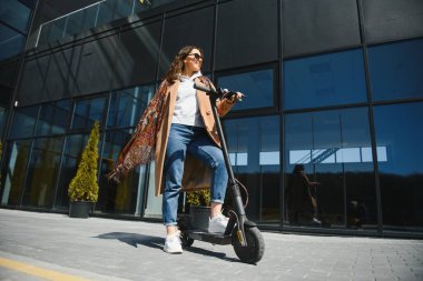 Genç ve güzel bir kız elektrikli scooterının yanında duruyor. Gülümsüyor, iş eğitimli, arka planda modern mimari var..