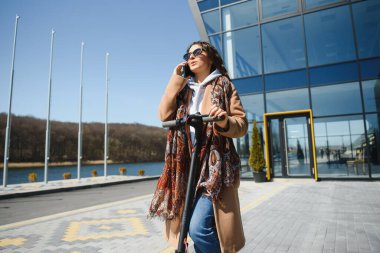 Ceketli genç ve güzel bir kadın ofis binalarında çalışırken gülümsüyor ve elektrikli scooter kullanıyor.