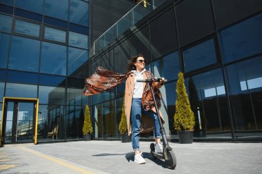 Genç ve güzel bir kız elektrikli scooterının yanında duruyor. Gülümsüyor, iş eğitimli, arka planda modern mimari var..