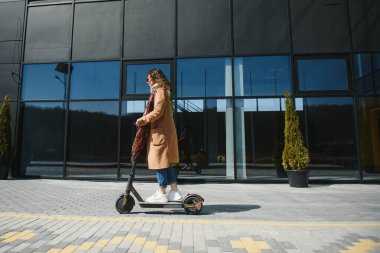 Genç ve güzel bir kız elektrikli scooterının yanında duruyor. Gülümsüyor, iş eğitimli, arka planda modern mimari var..
