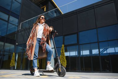 Ceketli genç ve güzel bir kadın ofis binalarında çalışırken gülümsüyor ve elektrikli scooter kullanıyor.