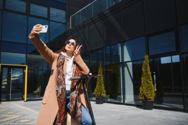 Genç ve güzel bir kız elektrikli scooterının yanında duruyor. Gülümsüyor, iş eğitimli, arka planda modern mimari var..