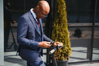 Büyük şehirde e-scooter sürerken telefonunu kontrol eden mutlu 30 'lu yaşlarda bir adam.