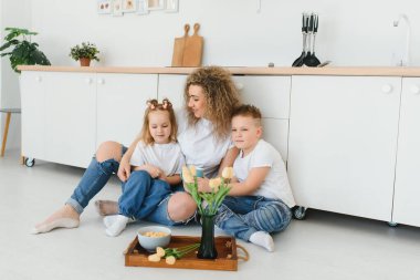 Mutlu genç bir aile, çocuklu bir anne yeni modern tasarım mutfağında sıcak ahşap zemine oturur, heyecanlı küçük çocukları olan bir anne dinlenir yenilenmiş bir dairede dinlenir, konsept taşınır.