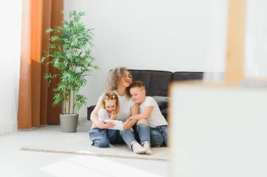 Güzel genç anne, küçük tatlı kızları ve oğulları tablet kullanıyorlar ve gülümsüyorlar, evde yerde oturuyorlar.