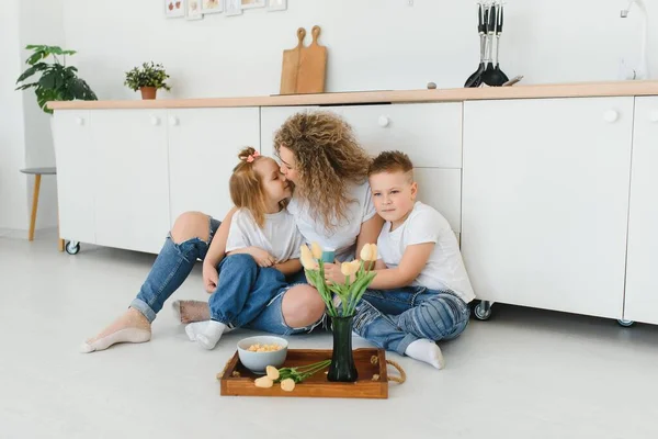 stock image Happy mother hugging daughter and son sitting on wooden floor in modern kitchen at home. Mom hugging children. Happy family relationship concept