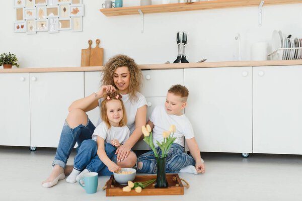 Happy mother hugging daughter and son sitting on wooden floor in modern kitchen at home. Mom hugging children. Happy family relationship concept