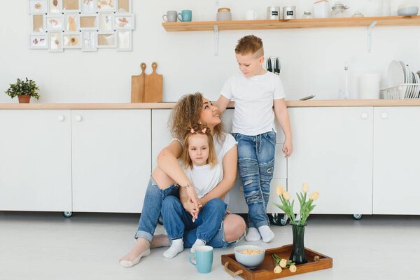 Happy young family, mom with little children sit on warm wooden floor in new modern design kitchen, mother with excited small kids relax rest in own renovated apartment, moving concept