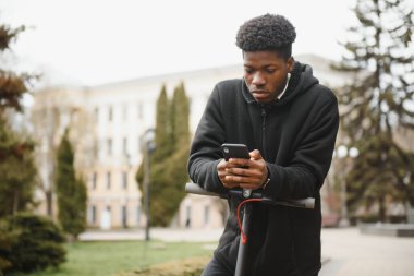 Afro-Amerikalı bir adam arka planda elektrikli scooter kullanıyor, bir öğrenci eko-taşıma kullanıyor.