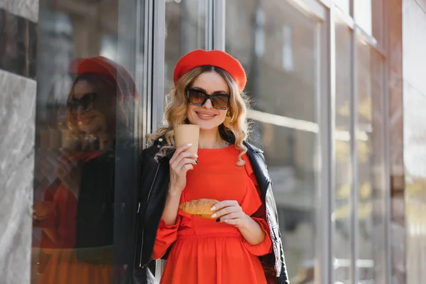 Pretty happy woman with sweet smile in vintage sunglasses in hat in casual red-black clothes with coffee walks and smiles outdoors in city. Cheerful urban fashion girl on walk in spring sunny day.