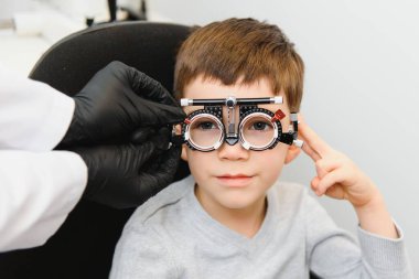 Küçük, ciddi bir çocuk görüş testi kürsüsünde oturuyor. Doktor özel gözlüklere lens takıyor.