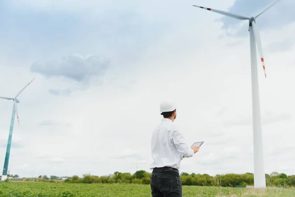 Engineer is checking energy production on wind turbine. Worker in windmills park in helmet and with tablet.