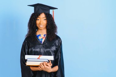 Siyah cüppeli ve şapkalı güzel bir Afro-Amerikalı kadın, mavi izole bir arka planda gülümsüyor.