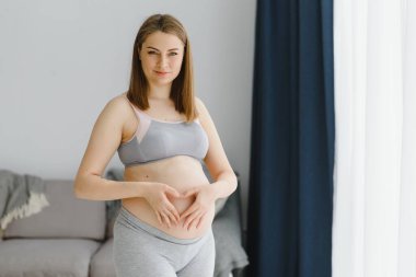 Güzel hamile kadın karnına dokunuyor ve evinin penceresinin yanında dururken gülümsüyor.