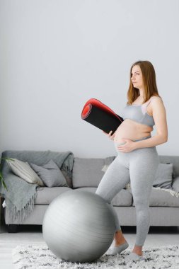 Spor kıyafetli gülümseyen hamile kadın sabah jimnastiğine ya da egzersize hazır. Mutlu genç kadın hamilelik boyunca sağlıklı yaşam tarzını izler, pilates veya esneme için yoga minderi tutar..
