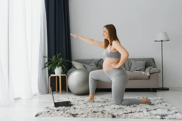 妊娠中の女性はラップトップで自宅でヨガを練習します 妊婦さんが室内で出産ビデオ研修をされています 女性の運動 妊娠中の瞑想 デジタルデバイス上のオンラインフィットネスクラス — ストック写真