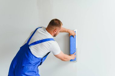 Sıvacı duvar yüzeyini bir duvar öğütücüsüyle düzleştiriyor. Usta inşaatçı beyaz alçı bir duvar öğütüyor. Tulumlu bir adam, yüzeyi solunum cihazıyla öğütür. deneyimli tamirci