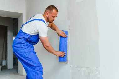 Tulumlu bir işçi duvarı çimentoyla kaplamak için bir mala kullanır.