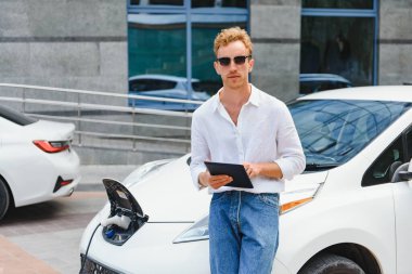 Gayri resmi giyinmiş genç bir adamın portresi, şarj istasyonunda duruyor. Eko elektrikli araba konsepti