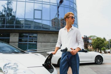 Gayri resmi giyinmiş genç bir adamın portresi, şarj istasyonunda duruyor. Eko elektrikli araba konsepti