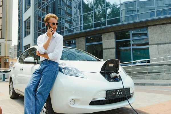 The guy sat down on the hood of the car. His car is charging at the charging station. A man looks at the smartphone screen and smiles.